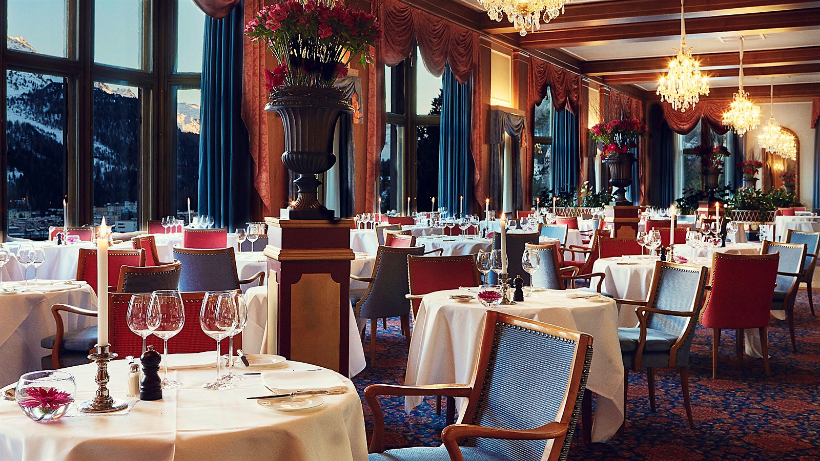 The dining room at Le Restaurant, with crystal chandeliers, chairs with blue-and-red seats, wood plinths holding black flower planters and windows offering mountain views